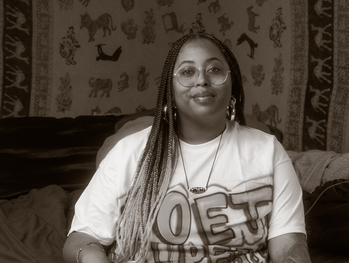 A nonbinary Black person with long braids fading to blonde, and round glasses. They are seated, wearing a t-shirt.