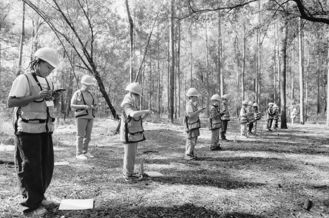 Young students in hardhats take measurements in the forest.