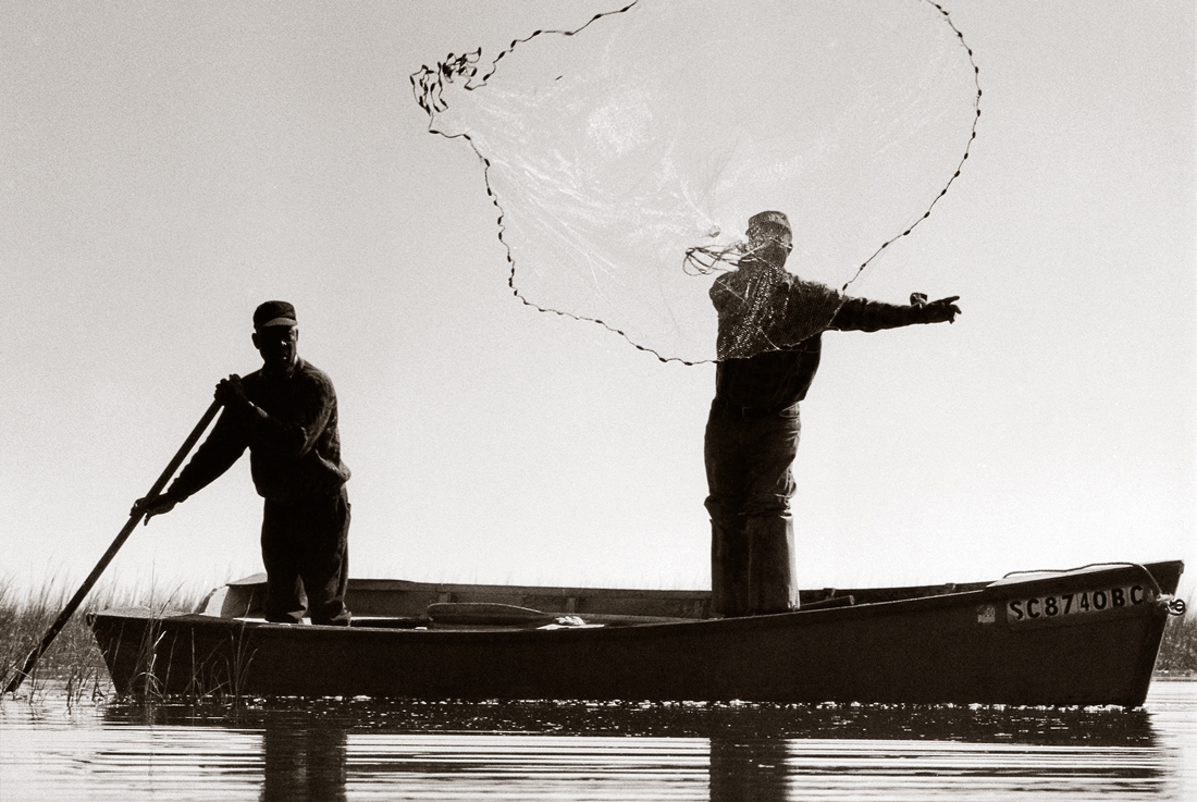 Two men in a small boat, one throws a net into the water.
