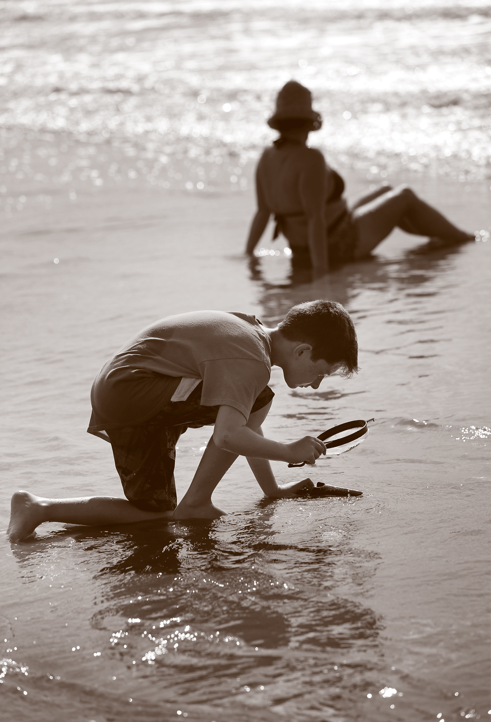 A young boy looks at the beach sand through a magnifying glass.