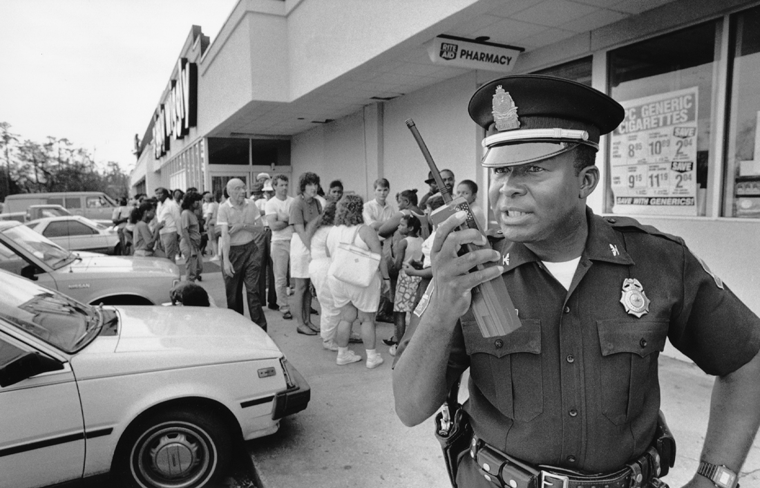 A police chief speaks into a walkie talkie in front of a line of people at a store.