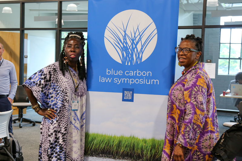 Two Black women in printed dresses pose in front of the Symposium sign.