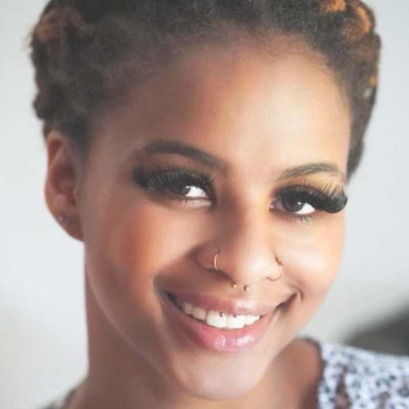 A smiling young Black woman with a nose piercing looks into the camera.