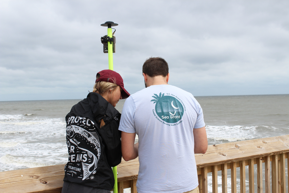 A man and woman hold a tall pole against the side of the pier looking out over the ocean.