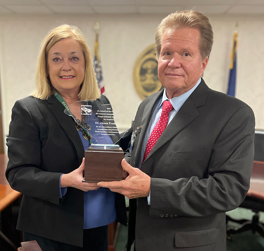 A middle aged white woman presents a glass award to a middle aged white man. Both are wearing formal attire in a government building.