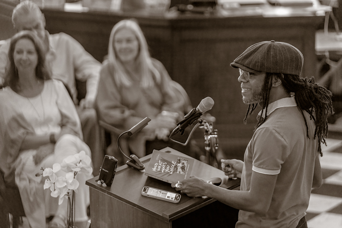 A Black man with dreadlocks and wearing a cap addresses a crowd from a podium.